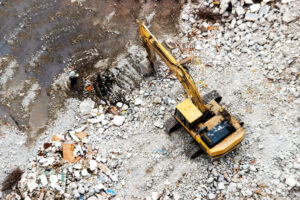 Vue aérienne d'une pelle mécanique jaune en cours d'excavation sur un chantier de dépollution, avec gravats, débris de démolition et zone humide sombre au sol, accompagnée d'un ouvrier en équipement de protection.