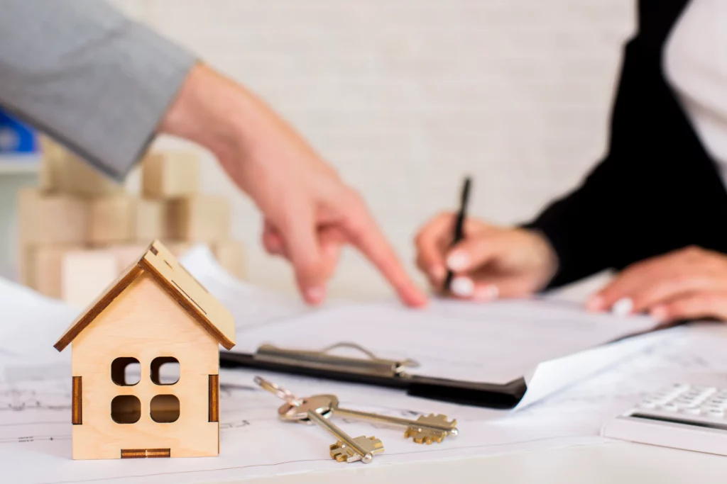 Maquette de maison en bois posée sur des plans immobiliers, avec un jeu de clés au premier plan et deux personnes signant un document en arrière-plan.